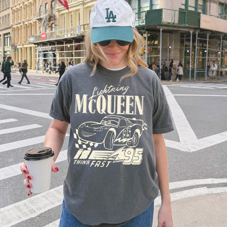 May include: A charcoal gray short-sleeve t-shirt featuring a graphic of the character Lightning McQueen with the text Lightning McQueen, 95, and Think Fast. The shirt is paired with a white and teal baseball cap with the letters LA in green.