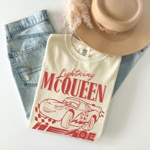 May include: A cream-colored t-shirt featuring a red graphic of the character Lightning McQueen with the text Lightning MCQUEEN and the number 95. The shirt is displayed alongside light blue denim jeans, a tan wide-brim hat, and a silver necklace.