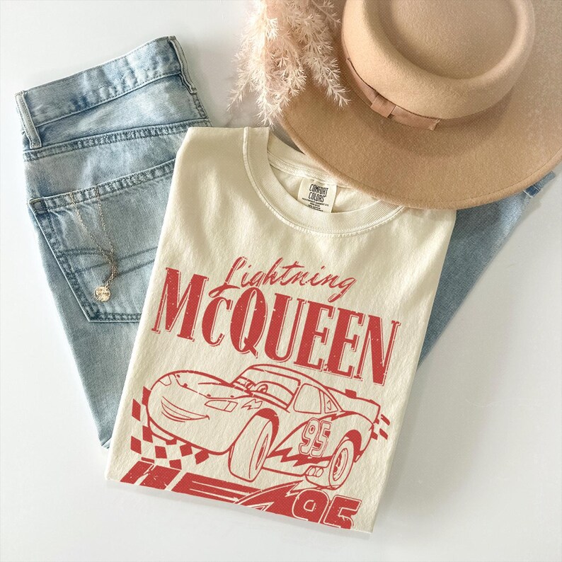 May include: A cream-colored t-shirt featuring a red graphic of the character Lightning McQueen with the text Lightning MCQUEEN and the number 95. The shirt is displayed alongside light blue denim jeans, a tan wide-brim hat, and a silver necklace.