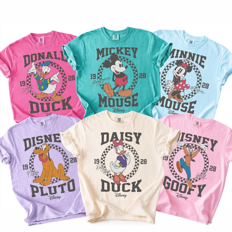 Op de afbeelding: Zes t-shirts met korte mouwen in roze, groenblauw, lichtblauw, lavendel, cr&egrave;me en lichtroze. Elk shirt heeft een Disney-personage, waaronder Donald Duck, Mickey Mouse, Minnie Mouse, Pluto, Daisy Duck en Goofy, met de tekst 1928.