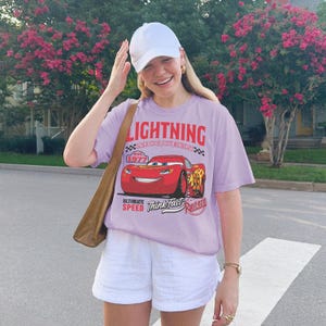 May include: A light purple short-sleeve t-shirt featuring a graphic of the red race car Lightning McQueen with the text Lightning McQueen, est. 1977, Ultimate Speed, Think Fast, and Rust-eze, paired with white shorts and a white baseball cap.