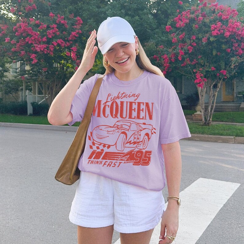 May include: A light purple short-sleeve t-shirt featuring a red graphic of the character Lightning McQueen with the text Lightning MCQUEEN and THINK FAST 95. The shirt is paired with a plain white baseball cap and white shorts.