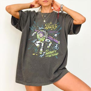 May include: A charcoal gray short-sleeve t-shirt featuring a graphic of Buzz Lightyear in flight. The design includes the text Buzz Lightyear and To Infinity and Beyond in yellow and blue, along with a small planet and a rocket ship emblem.