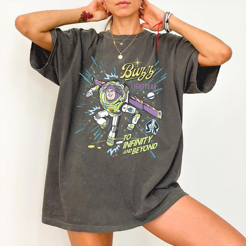 May include: A charcoal gray short-sleeve t-shirt featuring a graphic of Buzz Lightyear in flight. The design includes the text Buzz Lightyear and To Infinity and Beyond in yellow and blue, along with a small planet and a rocket ship emblem.