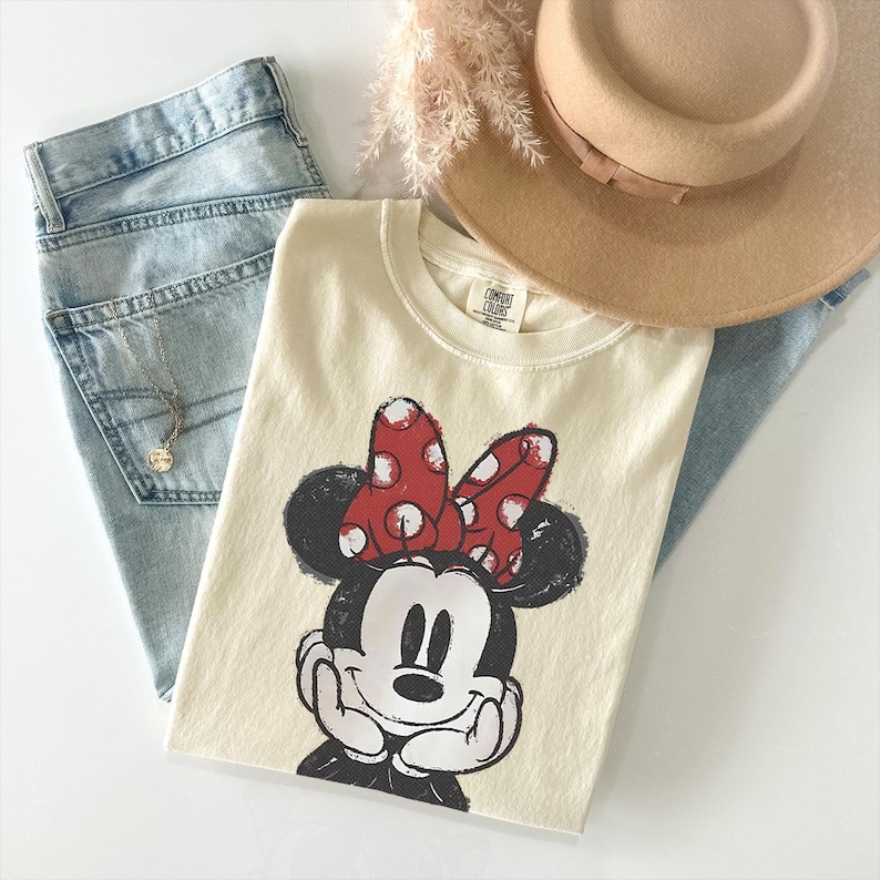Może przedstawiać: Kremowy t-shirt z grafiką Myszki Minnie w czerwonej kokardzie w białe kropki, zestawiony z jasnoniebieskimi dżinsami, beżowym kapeluszem z szerokim rondem oraz delikatnym złotym naszyjnikiem z małą okrągłą zawieszką.