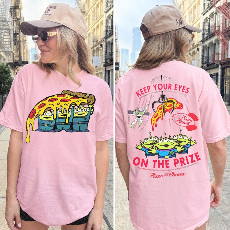 May include: A light pink t-shirt featuring a graphic of three green aliens holding a slice of pepperoni pizza on the front. The back displays a claw machine graphic with the text KEEP YOUR EYES ON THE PRIZE, a space ranger, and the Pizza Planet logo.