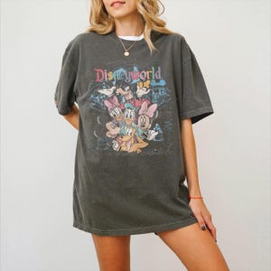 May include: A charcoal gray short-sleeve t-shirt featuring a graphic print of Disney characters including Mickey Mouse, Minnie Mouse, Donald Duck, Daisy Duck, Goofy, and Pluto. The text Disneyworld is printed in colorful letters above the characters.