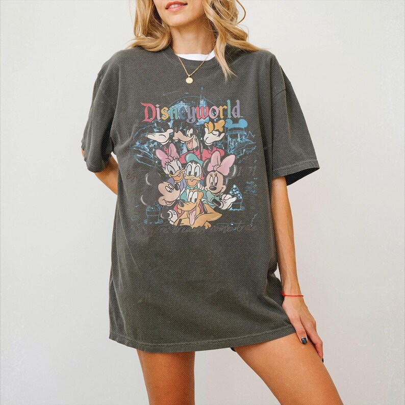 May include: A charcoal gray short-sleeve t-shirt featuring a graphic print of Disney characters including Mickey Mouse, Minnie Mouse, Donald Duck, Daisy Duck, Goofy, and Pluto. The text Disneyworld is printed in colorful letters above the characters.