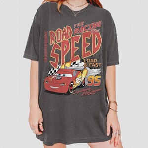 May include: A charcoal gray short-sleeve t-shirt featuring a graphic of a red race car with the text "THE ROAD RACING SPEED", "SUPERIOR PERFORMANCE", "LOAD & FAST", "95", and "Lightning McQueen" printed in red, yellow, and white colors.