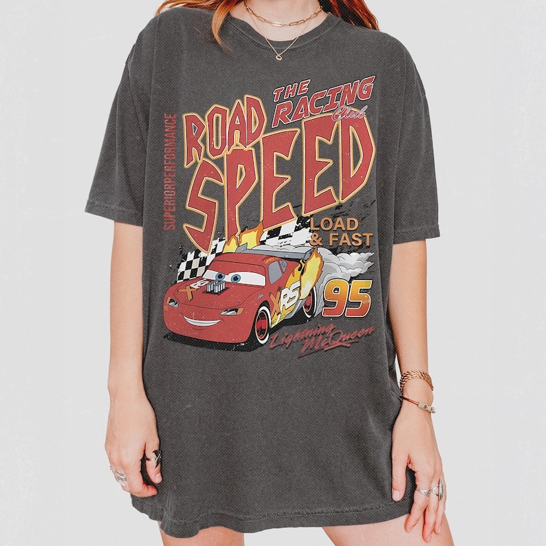 May include: A charcoal gray short-sleeve t-shirt featuring a graphic of a red race car with the text "THE ROAD RACING SPEED", "SUPERIOR PERFORMANCE", "LOAD & FAST", "95", and "Lightning McQueen" printed in red, yellow, and white colors.