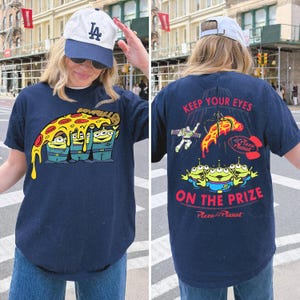 May include: A navy blue t-shirt featuring a graphic of three green aliens from Toy Story holding a slice of pepperoni pizza on the front. The back displays the text KEEP YOUR EYES ON THE PRIZE with Buzz Lightyear, the aliens, and a claw machine.
