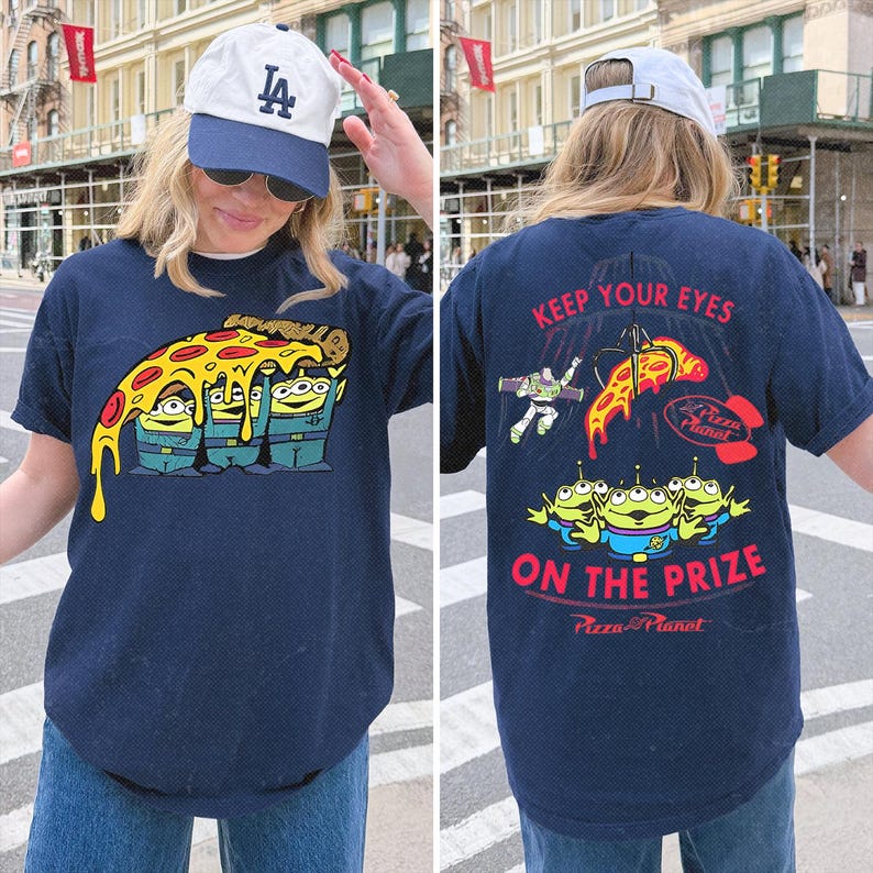 May include: A navy blue t-shirt featuring a graphic of three green aliens from Toy Story holding a slice of pepperoni pizza on the front. The back displays the text KEEP YOUR EYES ON THE PRIZE with Buzz Lightyear, the aliens, and a claw machine.