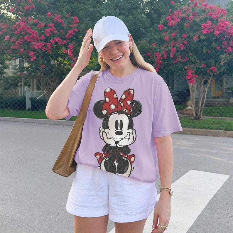 Może przedstawiać: Jasnofioletowy t-shirt z kr&oacute;tkim rękawem z nadrukiem graficznym przedstawiającym Myszkę Minnie w czerwonej kokardzie w białe kropki. Koszulka zestawiona jest z gładką białą czapką z daszkiem, białymi szortami i brązową torbą na ramię.