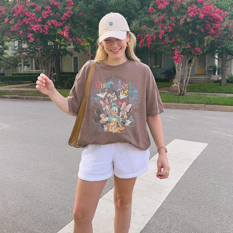 May include: A brown short-sleeved t-shirt featuring a colorful graphic print of Disney characters including Mickey Mouse, Minnie Mouse, Donald Duck, and Daisy Duck with the text Walt Disney World, paired with white shorts and a beige baseball cap.