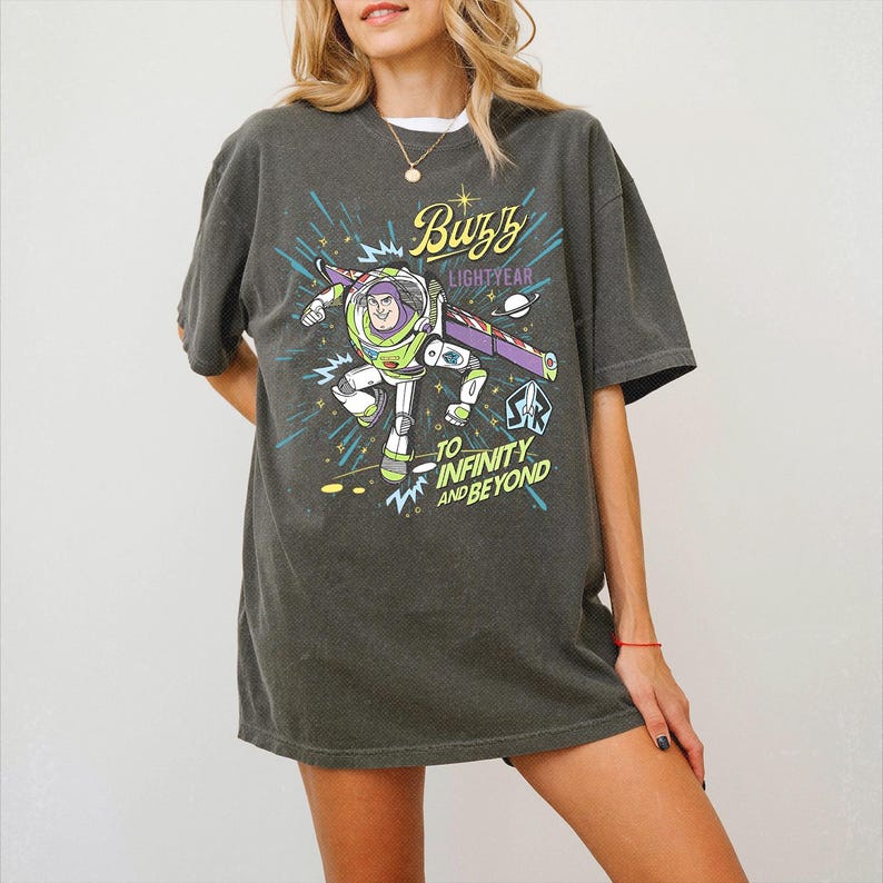 May include: A charcoal gray short-sleeve t-shirt featuring a graphic of Buzz Lightyear in a space suit with purple, green, and white accents. The text reads Buzz Lightyear and To Infinity and Beyond, accompanied by starbursts and a small planet graphic.