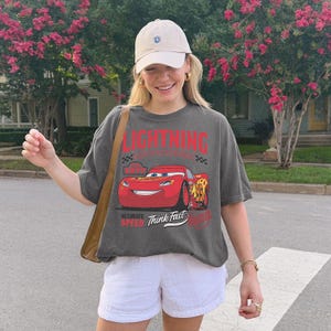 May include: A charcoal gray short-sleeve t-shirt featuring a graphic of a red race car with the number 95, the text LIGHTNING, 1977, ULTIMATE SPEED, and Think Fast. The shirt is paired with a beige baseball cap and white shorts.