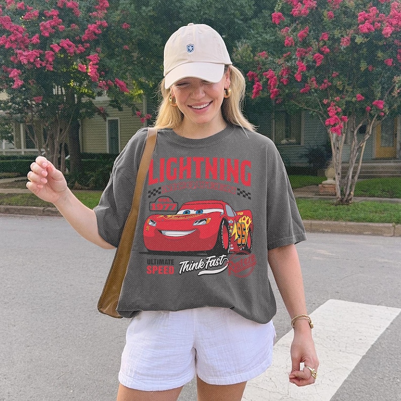 May include: A charcoal gray short-sleeve t-shirt featuring a graphic of a red race car with the number 95, the text LIGHTNING, 1977, ULTIMATE SPEED, and Think Fast. The shirt is paired with a beige baseball cap and white shorts.
