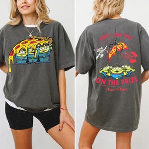 May include: A charcoal gray short-sleeve t-shirt featuring a graphic of three green aliens holding a slice of pizza on the front. The back displays the same aliens, Buzz Lightyear, a claw, and the text KEEP YOUR EYES ON THE PRIZE.
