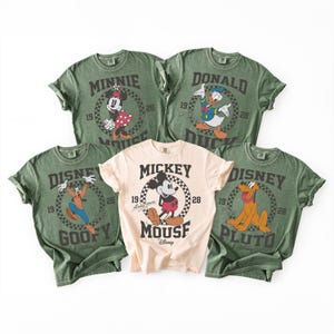 Op de afbeelding: Vijf T-shirts met korte mouwen en ronde hals met Disney-figuren. Vier shirts zijn olijfgroen met afbeeldingen van Minnie Mouse, Donald Duck, Goofy en Pluto. E&eacute;n centraal shirt is cr&egrave;me met een Mickey Mouse-afbeelding en tekst.