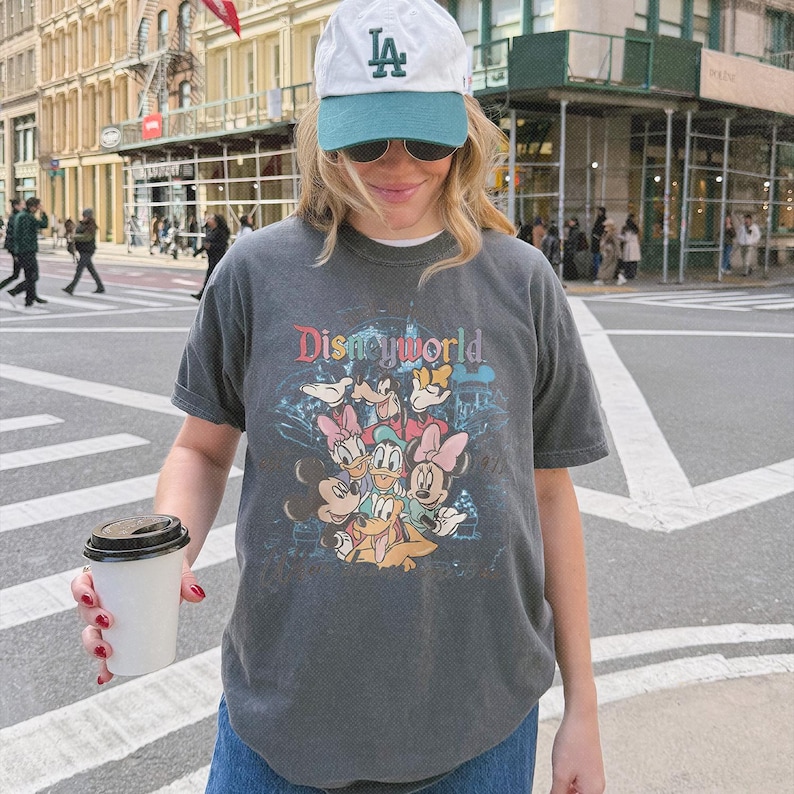May include: A charcoal gray short-sleeve t-shirt featuring a colorful graphic print of Disney characters including Mickey Mouse, Minnie Mouse, Donald Duck, Daisy Duck, and Pluto with the text Disneyworld above them, paired with a white and teal baseball cap.