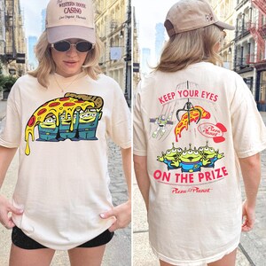 May include: A cream-colored t-shirt featuring a graphic of three green aliens under a slice of pizza on the front. The back displays a claw machine graphic with the text KEEP YOUR EYES ON THE PRIZE and the Pizza Planet logo in red and black.
