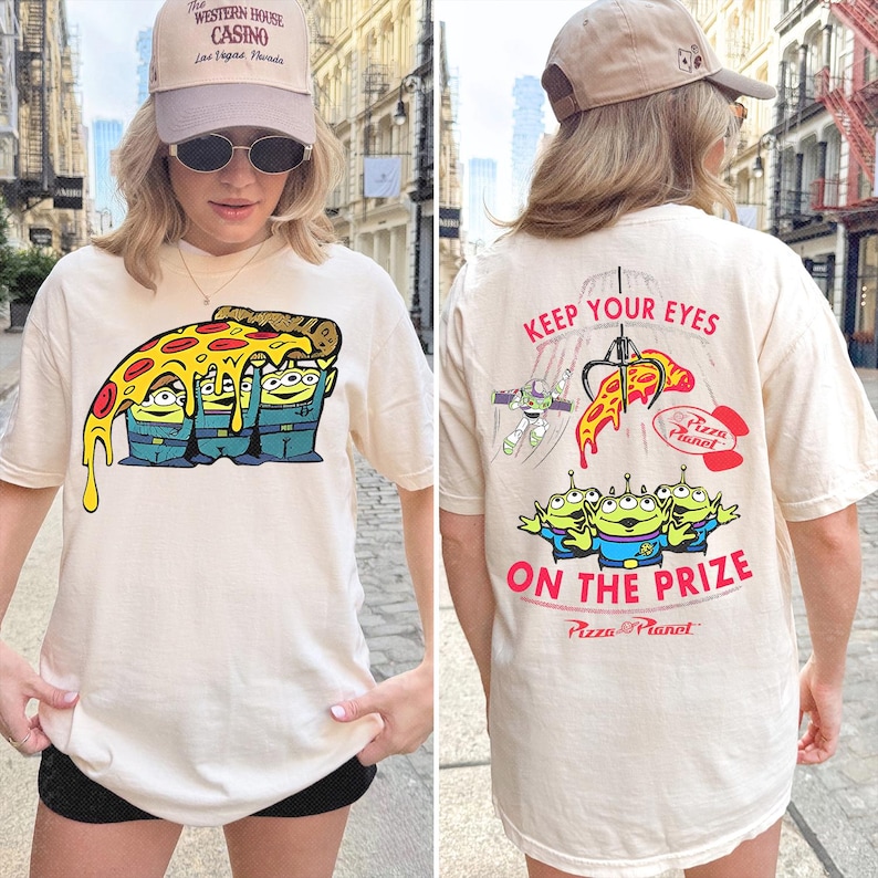 May include: A cream-colored t-shirt featuring a graphic of three green aliens under a slice of pizza on the front. The back displays a claw machine graphic with the text KEEP YOUR EYES ON THE PRIZE and the Pizza Planet logo in red and black.