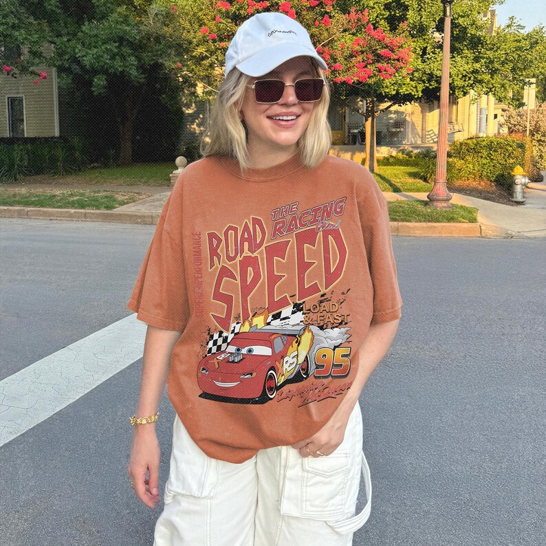 May include: A rust-colored short-sleeve t-shirt featuring a graphic of a red race car with the number 95 and the text ROAD RACING SPEED LOAD & FAST. The shirt is paired with a white baseball cap and white cargo pants with visible pocket details.