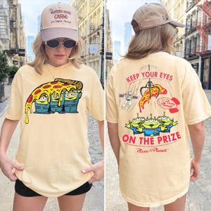 May include: A light yellow t-shirt featuring a graphic of three green aliens under a slice of pizza on the front. The back displays a claw machine graphic with the text "KEEP YOUR EYES ON THE PRIZE" and "Pizza Planet" above the aliens.