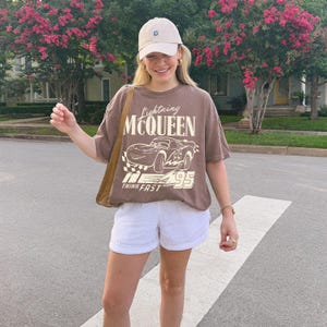 May include: A brown short-sleeve t-shirt featuring a graphic of the character Lightning McQueen with the text Lightning MCQUEEN and THINK FAST 95. The shirt is paired with white shorts, a beige baseball cap, and a brown shoulder bag.