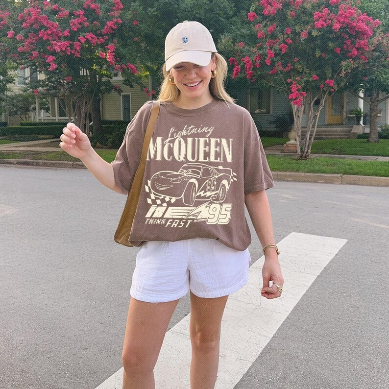 May include: A brown short-sleeve t-shirt featuring a graphic of the character Lightning McQueen with the text Lightning MCQUEEN and THINK FAST 95. The shirt is paired with white shorts, a beige baseball cap, and a brown shoulder bag.