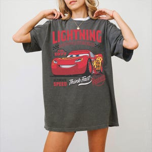 May include: A charcoal grey oversized t-shirt featuring a graphic of the red race car Lightning McQueen. The print includes the text LIGHTNING, 1977, ULTIMATE SPEED, Think Fast, and Rust-eze. The design has a distressed, vintage-inspired aesthetic.