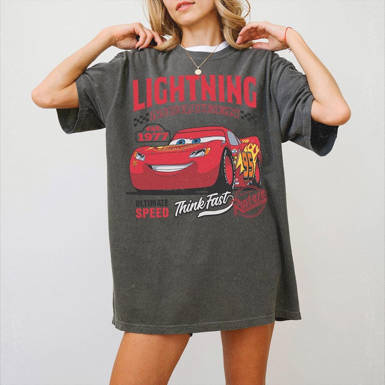 May include: A charcoal grey oversized t-shirt featuring a graphic of the red race car Lightning McQueen. The print includes the text LIGHTNING, 1977, ULTIMATE SPEED, Think Fast, and Rust-eze. The design has a distressed, vintage-inspired aesthetic.