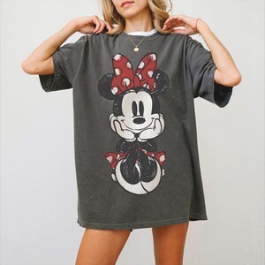 Może przedstawiać: Ciemnoszary t-shirt z kr&oacute;tkim rękawem z dużym, postarzanym nadrukiem graficznym przedstawiającym Myszkę Minnie. Postać ma na głowie czerwoną kokardę w białe kropki, pasującą sukienkę i opiera brodę na dłoniach w charakterystycznej pozie.