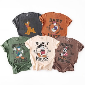 Op de afbeelding: Vijf T-shirts met korte mouwen en ronde hals in antraciet, oranje, groen, cr&egrave;me en bruin. Elk shirt is voorzien van een vintage-stijl afbeelding van een Disney-personage, waaronder Pluto, Daisy Duck, Donald Duck, Mickey Mouse en Minnie Mouse.
