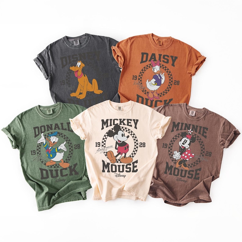 Op de afbeelding: Vijf T-shirts met korte mouwen en ronde hals in antraciet, oranje, groen, cr&egrave;me en bruin. Elk shirt is voorzien van een vintage-stijl afbeelding van een Disney-personage, waaronder Pluto, Daisy Duck, Donald Duck, Mickey Mouse en Minnie Mouse.