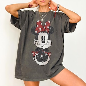 Może przedstawiać: Ciemnoszary t-shirt z kr&oacute;tkim rękawem z nadrukiem graficznym w stylu vintage przedstawiającym Myszkę Minnie z przodu. Postać ma na sobie charakterystyczną czerwoną kokardę w białe kropki, czarne uszy i białe rękawiczki, opierając podbr&oacute;dek na dłoniach.