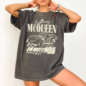 May include: A charcoal gray short-sleeve t-shirt featuring a graphic of the character Lightning McQueen. The print includes the text Lightning MCQUEEN, the number 95, and the phrase THINK FAST in a distressed white font with a checkered pattern.