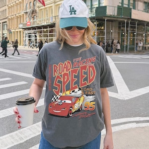 May include: A charcoal gray short-sleeve t-shirt featuring a graphic of a red race car with the text THE ROAD RACING SPEED LOAD & FAST 95 LIGHTNING MCQUEEN. The shirt is paired with a white and teal baseball cap with the letters LA in green.