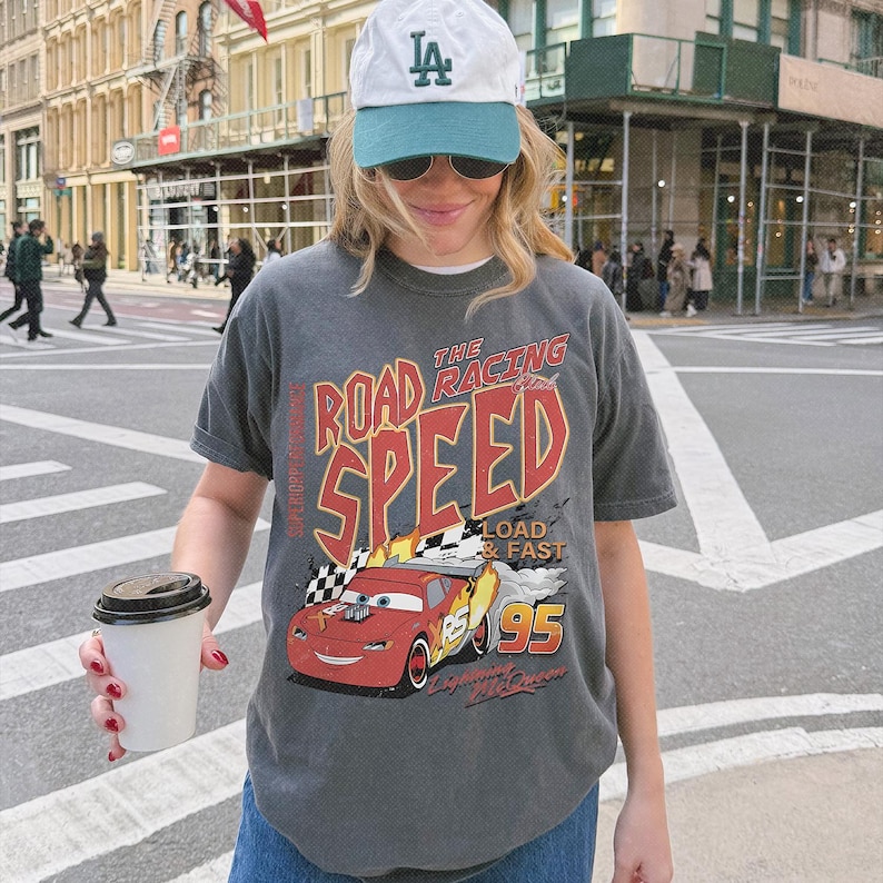 May include: A charcoal gray short-sleeve t-shirt featuring a graphic of a red race car with the text THE ROAD RACING SPEED LOAD & FAST 95 LIGHTNING MCQUEEN. The shirt is paired with a white and teal baseball cap with the letters LA in green.