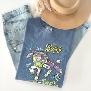 May include: A blue crew neck t-shirt featuring a graphic of Buzz Lightyear in a space suit with the text Buzz Lightyear To Infinity And Beyond, accompanied by light blue denim jeans, a tan wide-brimmed hat, and a delicate gold chain necklace.