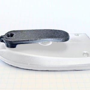 May include: A black, textured, rectangular object with a curved handle, resting on a white, rounded base. The base has a smooth surface with a few small, circular indentations. The object appears to be a component of a larger device.