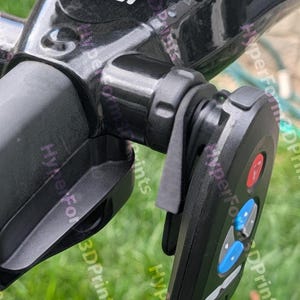 May include: Close-up of a black device with the letters "MGI" in white. The device has a control panel with red, blue, and gray buttons. The background is blurred green grass.
