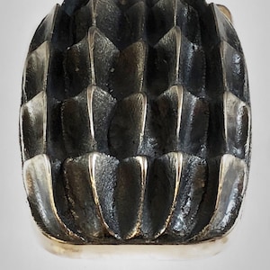 May include: A silver ring with a textured, geometric design resembling a pineapple. The ring is made of metal and has a shiny finish.
