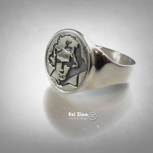 Op de afbeelding: Een zilveren ring met een rond, gegraveerd ontwerp met een portret van een persoon. De ring ligt op een witte achtergrond.