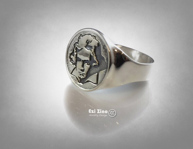 Op de afbeelding: Een zilveren ring met een rond, gegraveerd ontwerp met een portret van een persoon. De ring ligt op een witte achtergrond.