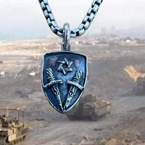 Peut inclure: Un collier en argent avec un pendentif en forme de bouclier. Le bouclier présente une étoile de David et deux épées croisées. Le pendentif est suspendu sur un fond flou de véhicules militaires.