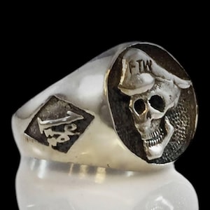 May include: Silver ring featuring a skull design on a textured background. The ring has a diamond-shaped emblem with an inscription and the letters "FTW" on the skull's hat. The ring is set against a dark background.