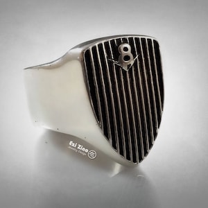Puede incluir: Un anillo de plata con un diseño en forma de escudo que presenta un patrón de rayas blancas y negras y el número "8" en el centro.