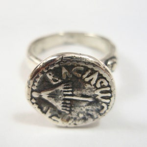 May include: A silver ring with a round, textured top featuring an inscription that reads "IN A.C.U.S."