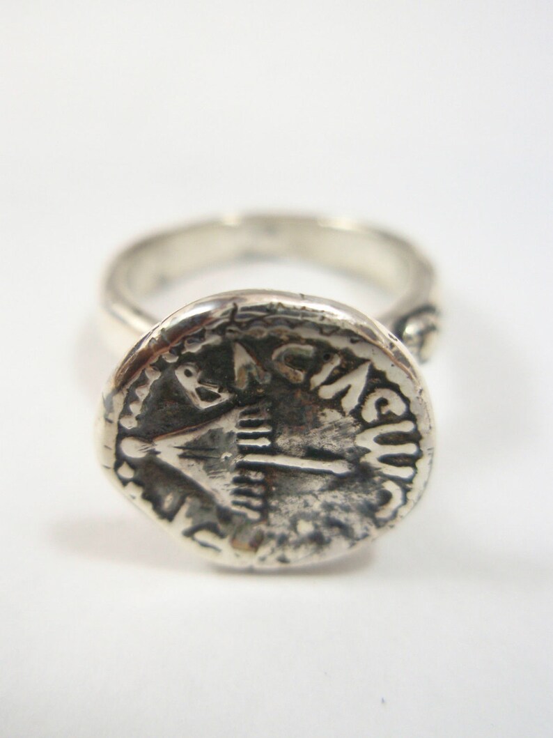 May include: A silver ring with a round, textured top featuring an inscription that reads "IN A.C.U.S."